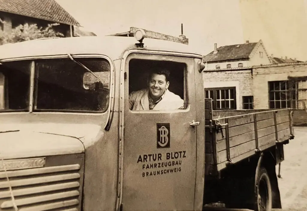 Schwarz-wei&szlig;-Fotografie eines historischen Lastwagens der Marke B&uuml;ssing-NAG mit einem l&auml;chelnden Fahrer im Fahrerhaus. Auf der Fahrert&uuml;r befindet sich das Logo und der Schriftzug &bdquo;Artur Blotz Fahrzeugbau Braunschweig&ldquo;.