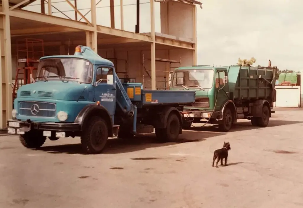 Historisches Farbfoto auf dem Betriebshof von Bl&ouml;tz. Im Vordergrund ein blauer Mercedes-Benz Kurzhauber-Lastwagen mit Pritsche und gelber Rundumleuchte, dahinter ein gr&uuml;ner Mercedes-Benz Frontlenker-Lkw mit M&uuml;llcontainerschlitten. Der Hintergrund zeigt eine offene, st&auml;hlerne Lagerhalle im Bau.