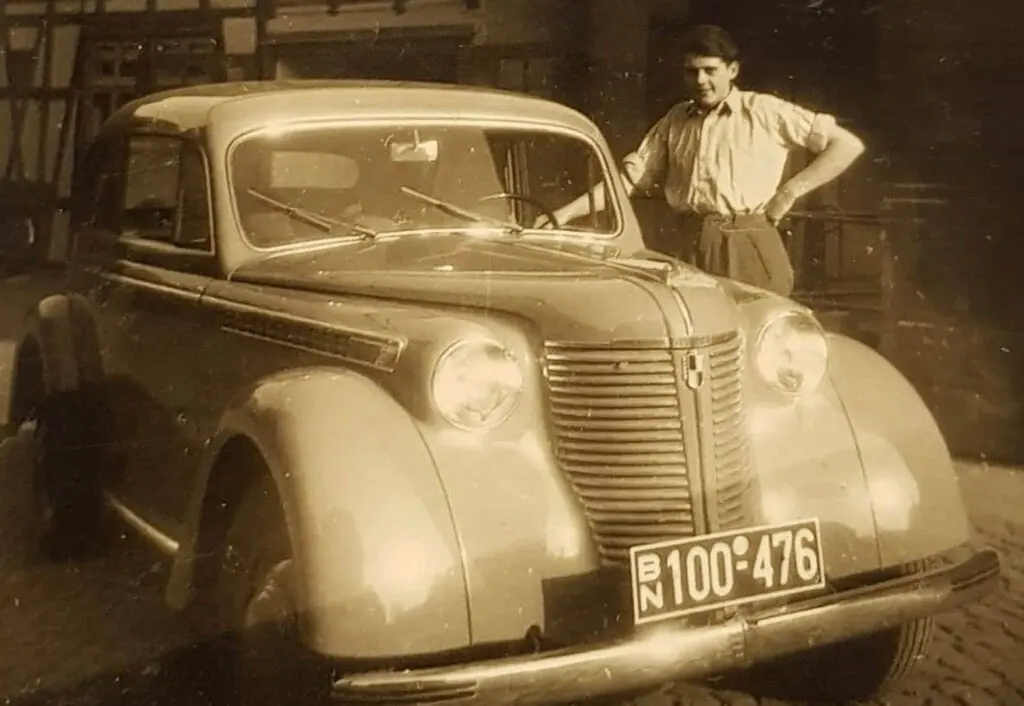 Eine historische Schwarz-Wei&szlig;-Fotografie (Sepia) eines klassischen PKW aus den 1940er oder fr&uuml;hen 1950er Jahren. Neben dem gl&auml;nzenden Wagen steht ein junger Mann in zeitgen&ouml;ssischer Kleidung mit einer Hand an der Fahrert&uuml;r.